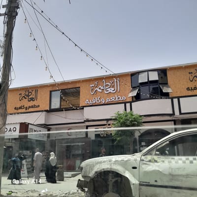 The image shows a street scene in Nuseirat, Palestinian Territories. A building with signs in Arabic advertising "Al-Azama Restaurant and Cafe" ("El Aazma" and "Al-Azama") is visible. There is a damaged white car with "PUMA" written on the side next to the building. People in traditional Islamic attire are walking on the sidewalk, and there is an advertisement for "MADAV NUTS, COFFEE & MORE" in the background. The image was taken with a device bearing a "Galaxy A14" watermark. String lights are strung overhead.
