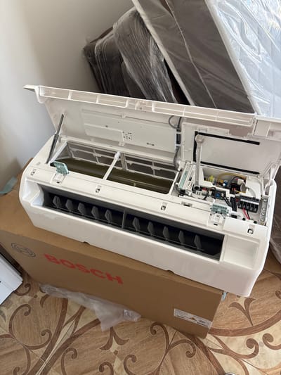 An indoor unit of a Bosch air conditioner is shown, its front cover open, revealing internal components. The unit is placed on top of a Bosch branded cardboard box. In the background, two mattresses wrapped in plastic are leaning against a wall. The scene appears to be indoors, possibly a room under renovation or storage area, with tiled flooring visible. The overall impression is of a newly acquired appliance. The image does not provide any specific clues about its location in Mezdreya, Bulgaria.