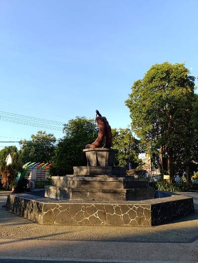 A stone fountain with a brown abstract sculpture on top, situated in a park in Kecamatan Asembagus, Indonesia, with coordinates -7.75 latitude and 114.218 longitude. The fountain is made of dark grey stone, with a square base and a layered design, rising to the top where the sculpture is placed. There are two people sitting in front of the fountain, with their backs to the viewer. The fountain is in the center of the scene, surrounded by green trees. The sky is clear blue and the sun casts long shadows on the ground. The park is clean and well maintained.