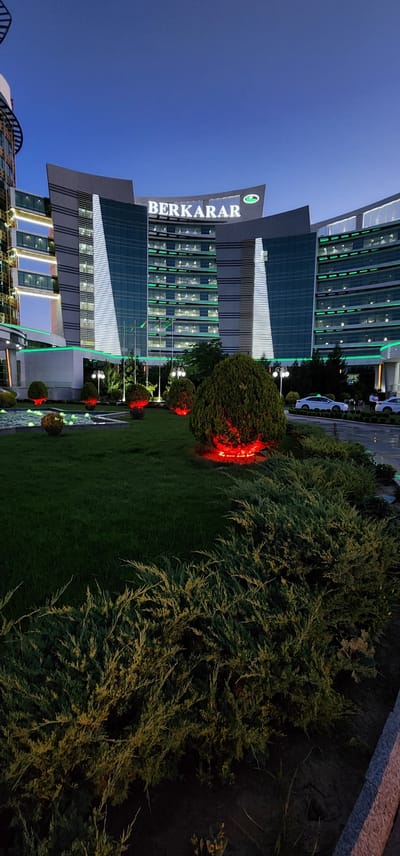 The image shows a modern building complex in Türkmenbaşy, Turkmenistan, at dusk. The architecture is characterized by sleek lines and a prominent "BERKARAR" logo illuminated in white on one of the buildings. The building's facade is adorned with green accent lighting, giving it a futuristic and sophisticated appearance. In the foreground, a well-maintained garden area features manicured bushes and trees, some of which are illuminated with red lights, creating a striking contrast against the twilight sky. The scene suggests a commercial or business area, possibly a corporate headquarters or a shopping center, given the grandeur of the buildings. The time of day is evident from the deep blue of the sky transitioning into darkness, indicating early evening. The weather appears calm, with no signs of precipitation. The overall ambiance is one of modern development and aesthetic design, with the vibrant lighting adding a touch of drama to the urban landscape. No people are clearly visible in the image, allowing the focus to remain on the architecture and landscaping.