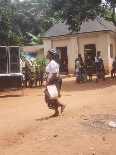 The image depicts an outdoor scene in Umundugba, Nigeria. A woman is walking on a sandy surface. She is wearing a white top, a dark patterned skirt, a head wrap, and sandals, and carrying a pink bag. A stack of colorful plastic chairs is visible behind her, along with several large speakers. In the background, there is a building with beige walls, some individuals standing in front of it, and a motorcycle parked nearby. A tree with lush green foliage is on the left side of the frame.
