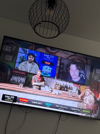 The image shows a television screen displaying a broadcast of the ESL Cologne Major 2026 esports event. On the screen, there are two individuals, one is a man with a beard wearing a light-colored shirt and headphones, positioned in the upper left part of the screen. To the right, another man with wavy hair is visible. In the foreground of the broadcast, a figure wearing a red bandana and a brown jacket is standing behind a counter. The background of the broadcast appears to be a virtual bar setting. Below the broadcast, a series of sponsor logos are displayed, including Intel, ESL, Betking, Maincast, Olx, Visa, PrivatBank, Logitech, and Okko. The television is placed in a room, with a black, wire-frame chandelier hanging from the ceiling above it. The image is taken from a slightly elevated angle, looking down at the television. The specific location within the city of Gothenburg, Sweden, cannot be determined from the provided image.