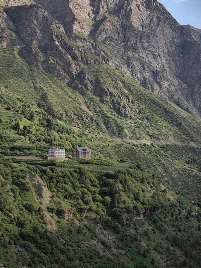 The image depicts a mountainous landscape with two buildings situated on a terraced hillside. The terrain is steep and covered in lush green vegetation, including scattered trees and shrubs. The buildings appear to be residential, with multiple stories and distinct architectural features. One building is lighter in color with numerous windows, while the other is darker with a prominent roofline. A narrow, winding road is visible in the middle ground, ascending the mountain. The background is dominated by a massive, rocky mountain range, exhibiting layers of geological formations. The scene is located in Kardang, India, a region known for its picturesque mountainous terrain and villages nestled within valleys. The lighting suggests it is daytime, with shadows cast across the landscape, indicating a natural, outdoor environment.