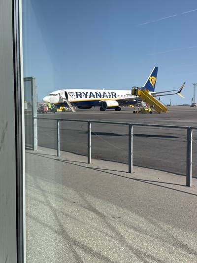 The media file depicts a Ryanair airplane parked on an tarmac, likely at an airport. The plane's livery is white with blue and yellow accents, and the word "RYANAIR" is prominently displayed along its fuselage in large blue lettering. A yellow passenger boarding stairs is attached to the aircraft's door. Several airport vehicles, including what appear to be baggage tugs or service carts, are visible near the plane.
The scene is set outdoors under a clear, bright blue sky, suggesting a sunny day, possibly in the morning or afternoon due to the strong shadows cast on the tarmac. The tarmac itself appears to be asphalt, with lines and markings indicating its function as a service area. In the foreground, a reflective surface, possibly a window, partially frames the image, creating a sense of looking out from a terminal building.
The overall atmosphere is one of typical airport operations, with an aircraft preparing for or having just completed a flight. There are no visible people clearly identifiable as distinct individuals, but the presence of airport vehicles implies human activity related to the aircraft's servicing. The location context is Kastrup, Denmark. No specific emotions are conveyed, and the focus is on the industrial and functional aspect of an airport environment.