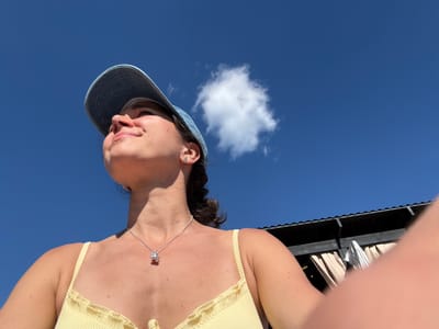 A woman wearing a light yellow ribbed bikini top and a light blue denim baseball cap looks upwards. A silver necklace with a pendant is visible on her neck. She has a slight smile. The background is a clear blue sky with a single white cloud and a portion of a dark wooden structure with draped fabric and a striped towel. The scene is outdoors, possibly in a sunny location in or near the city of Рыбхоз, Russia.