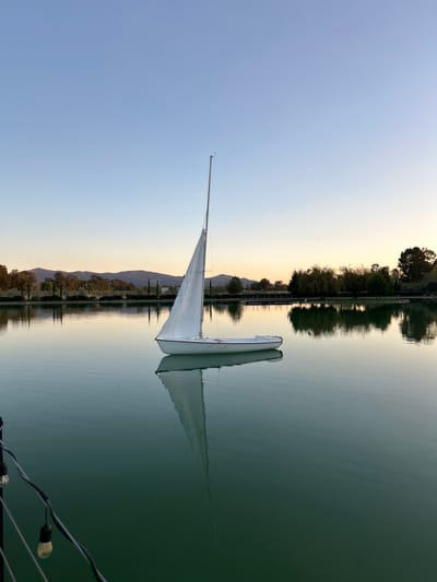 A sailboat sits in calm water reflecting the surrounding landscape. A long, thin sail extends upwards towards a blue sky. Behind the boat, a row of trees and mountains fades into the horizon. The water reflects the sky and trees, creating a symmetrical composition. In the lower right corner, a string of lights adds a touch of artificial illumination. The scene appears tranquil and serene.