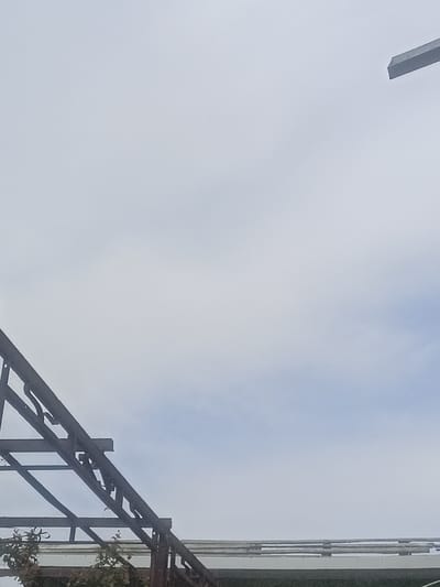 The image shows an overcast sky with a few wisps of clouds. In the foreground, there is a dark, angular metal structure, possibly part of a greenhouse or a similar construction, that extends from the left side of the frame upwards and to the right. Some leafy green foliage is visible at the bottom left, peeking out from behind the metal framework. The overall impression is of an outdoor scene, likely during daylight hours given the sky conditions, with a focus on the industrial or agricultural structure against the muted backdrop of the sky. There is a sharp, angled metallic object in the upper right corner, which could be part of another structure or an overhead fixture. The weather appears to be cloudy, creating a soft, diffused light. No people are visible in the image.
