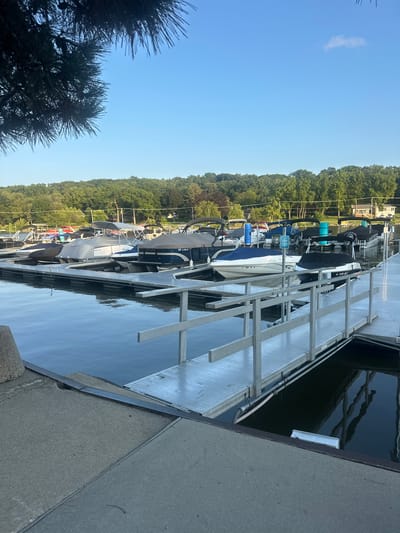 The image shows a marina with several boats docked on a calm body of water. The boats are covered with tarps, except for a few that are partially visible. There is a wooden dock with a metal railing extending into the water, providing access to the boats. In the background, there is a shoreline covered with trees and some houses. The sky is clear and blue, with a few white clouds. The overall scene suggests a peaceful and quiet morning at the marina.