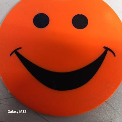 The image shows a close-up of an orange smiley face, likely a sticker or a decoration, placed on a textured, light-colored surface. The smiley face is depicted with two black circular eyes and a wide black smile. The lighting is even, highlighting the texture of the orange surface. In the bottom left corner of the image, the text "Galaxy M32" is visible, indicating the device used to capture the photo. The scene appears to be an indoor setting, possibly a desk or table. There is no indication of the city of Olsztyn, Poland in the image.