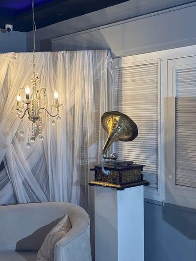 The image captures an indoor scene, likely a room with a vintage aesthetic, located in Sweileh, Jordan. In the foreground, a plush, light-colored sofa is partially visible on the left. To the right, a vintage gramophone with a brass horn and ornate wooden base sits atop a white pedestal. The gramophone is positioned in front of a window with white louvered blinds. Behind the gramophone and sofa, sheer white drapes create a soft, diffused backdrop. A crystal chandelier with multiple lights hangs from the ceiling above the drapes. A security camera is visible in the upper left corner of the image, mounted on the ceiling. The overall atmosphere suggests a decorative display or a room designed for ambiance.