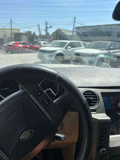 An interior shot of a Land Rover vehicle featuring the steering wheel prominently. The steering wheel has control buttons and a Land Rover logo. The dashboard with air vents and other controls is partially visible. Outside the vehicle, a row of cars, mostly white, is seen parked on a street with buildings and power lines in the background under a clear blue sky. The view is slightly obstructed by a dirty windshield.