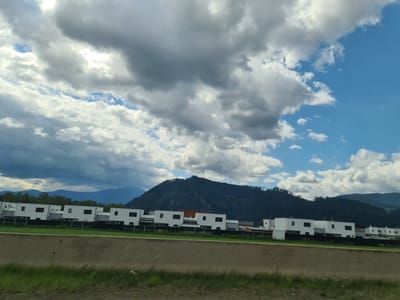 This image captures a modern residential area in the city of Judenburg, Austria, under a dynamic sky filled with dramatic clouds and patches of blue. In the foreground, a concrete barrier separates the view from the residential buildings. The buildings themselves are contemporary, uniformly white with dark rectangular windows, arranged in a row. Behind the houses, a strip of green grass is visible, followed by a dark fence. The backdrop consists of lush green, tree-covered mountains that rise majestically, their peaks partially obscured by the sky and lower-lying clouds. The overall impression is one of a peaceful, suburban landscape juxtaposed with imposing natural scenery, all under a moody, cloud-filled sky.