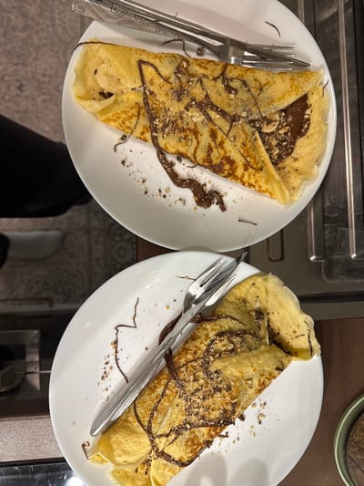The image displays two white plates each holding a generously filled crepe, likely prepared in Maldegem, Belgium. The crepes are golden-brown and folded, with their interiors revealing a rich, dark chocolate spread, possibly Nutella. Both crepes are sprinkled with a granular topping, appearing to be crushed nuts or cookies, and feature decorative drizzles of the same chocolate spread. One of the crepes clearly shows slices of banana tucked inside, adding another layer to the filling.
Each plate is accompanied by a silver fork, placed beside the crepe, suggesting they are ready to be eaten. The plates rest on what appears to be a dark-colored kitchen counter or table. In the background, hints of a dark appliance or countertop are visible on the left, and a green-rimmed jar is partially seen on the right. Below the plates, the flooring has a subtle patterned texture, and a person's legs or feet, clad in dark clothing and light shoes, are partially visible, indicating an immediate presence in the scene. The overall presentation suggests a home-cooked meal, possibly breakfast, a snack, or dessert, prepared with care. No visible text is present in the image.
