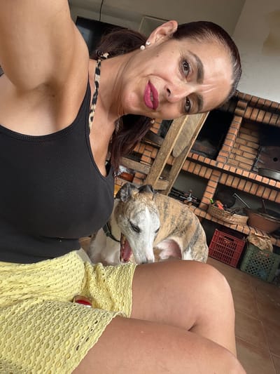 A woman with dark hair pulled back into a ponytail and wearing a black tank top and a yellow eyelet skirt is taking a selfie. She has bold pink lipstick on and is wearing earrings. She is sitting next to a greyhound with a brindle coat that is panting. In the background, there is a brick barbecue grill, a wooden structure, and plastic crates. The scene appears to be outdoors on a tiled floor. This image was captured in the city of Caldas da Rainha, Portugal.