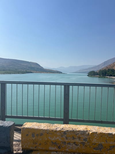 The image depicts a scenic view of a large body of turquoise water, likely a reservoir or lake, surrounded by rolling, arid hills. In the foreground, a grey metal railing runs horizontally across the frame, obscuring the lower portion of the water. Beyond the railing, two weathered concrete barriers are visible on the left side of the frame, one grey and the other painted yellow and white, showing signs of age and wear. The water's surface is calm, with subtle ripples reflecting the clear blue sky above. Distant hills and mountains, some sparsely covered with vegetation, frame the expansive water. On the right side of the water, a line of trees suggests a developed area, possibly a park or settlement. The overall impression is one of a tranquil, natural landscape under a bright, clear sky. Based on the prompt, this scene is located in the city of Yanghikurgan, Uzbekistan.
