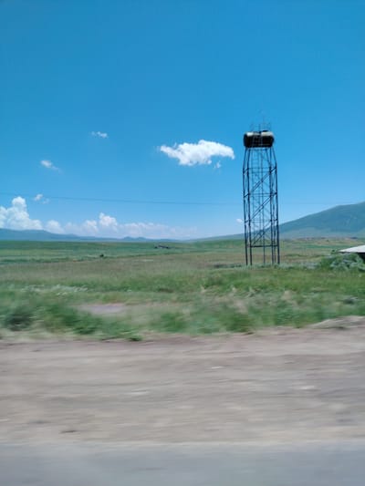 In the city of Hartavan, Armenia, under a clear blue sky with scattered white clouds, a tall, dark metal water tower stands prominently in a vast, green field. The tower features a cylindrical tank at the top and a ladder system ascending its structure. The surrounding landscape is characterized by rolling hills in the distance and a blurred foreground suggesting motion, possibly from a vehicle. The scene is open and rural, with a sense of distance and expanse.