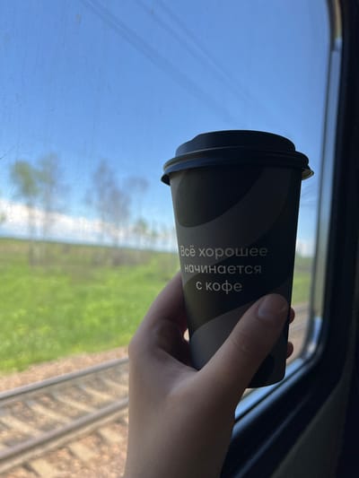 A hand holds a disposable coffee cup with text on it in front of a train window. The cup is dark gray with lighter gray wavy patterns. The text reads "Всё хорошее начинается с кофе" which translates to "Everything good starts with coffee." The window shows a blurry green landscape with trees and blue sky, suggesting the train is moving through a rural area. The train tracks are visible below the landscape. There is no visible indication of the specific city of Zoevka, Russia.
