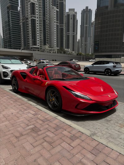 The image depicts an urban street scene in Business Bay, United Arab Emirates, during a bright, sunny day. The main subject is a vibrant red Ferrari convertible, likely a 488 Pista Spider or similar model, prominently parked on the side of a road. Its red leather interior and yellow brake calipers, visible through the dark gray wheels, are notable details.

Behind the Ferrari, a dark gray Polestar 2 SUV is parked, with "POLESTAR" visible on its rear. Further along the street, several other vehicles are parked, including a dark red sedan and a white SUV, which appears to be a Kia based on visible branding and a partially obscured window sticker showing "KIA" and contact details.

The background features numerous modern high-rise buildings with varied architectural styles, including one with horizontal louvers and another with large glass windows. Text visible on these buildings includes "SOHO," "Al Fardan," and "Al-Qudra," likely indicating business or residential names. The sidewalk is paved with red brick-patterned blocks. The bright sunlight casts distinct shadows, suggesting a time between late morning and early afternoon. No people or specific activities are visible other than the parked cars. The overall setting is a developed and affluent urban environment.
