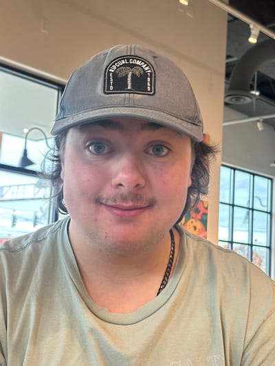 This image presents a frontal portrait of a young adult male situated within a cafe or similar establishment in Medford, United States. The subject, a light-skinned male with blue eyes and dark, medium-length hair visible at the sides, wears a faded gray baseball cap. The cap features a black rectangular patch embroidered with a white palm tree, 'RIPCURL COMPANY', 'ESTD', and '1969'. He has a visible mustache and a faint smile, with his eyes wide open. He is dressed in a light green, short-sleeved t-shirt with a worn collar and a dark, presumably metallic, chain necklace.
The background on the left features a large window, through which a light-colored building, a dark outdoor lamp fixture, and a partial sign displaying the capitalized letters 'VAU' are visible. To the right, another multi-paned window reveals an outdoor scene with bare tree branches. Between the subject and the right window, a colorful abstract art piece, characterized by orange and dark organic shapes, hangs on a wall. The ceiling exhibits exposed black ductwork and track lighting, consistent with a modern commercial interior.
