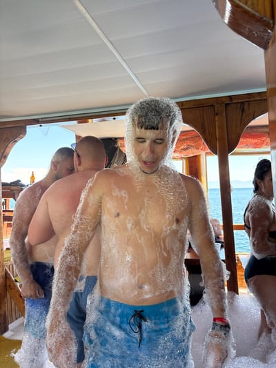 This image captures a vibrant foam party on a boat, likely off the coast of Menderes, Turkey. The primary subject is a young man, shirtless and wearing blue swim shorts, covered head-to-toe in white foam. He appears to be smiling with his eyes closed, suggesting enjoyment. His hair is also heavily coated in foam.

In the background, several other individuals are visible, similarly covered in foam. One person, partially obscured, wears a black top and is also covered in suds. Another man with a shaved head and sunglasses rests his arm on a wooden railing, also covered in foam, with another person behind him. A red wristband is visible on the arm of one person. The boat features wooden railings and structures, indicating a classic boat or gulet design. The ceiling above is white with horizontal panels.

Beyond the boat, the bright blue sea stretches out under a clear, sunny sky, with distant land or mountains visible on the horizon, confirming a coastal setting. The overall atmosphere suggests a lively and fun daytime event. There is no visible text in the image. A yellow bottle can be faintly seen in the reflection of the boat's polished surface in one of the cropped images.