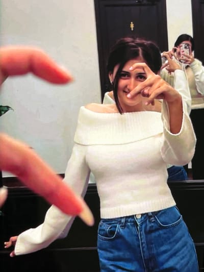 The image shows a young woman with dark hair, wearing a white off-the-shoulder sweater and blue jeans. She is in a room with dark walls and a mirror. In the mirror, another person is visible taking a selfie. The woman in the foreground is making a heart shape with her hands over her face. There are also two blurred hands in the foreground, one making a heart shape and another extended towards the woman. This appears to be a candid or posed photograph taken indoors, possibly in a restroom or a fitting room, given the presence of a mirror and the styling of the environment. There is no visual information in the image to suggest any connection to the city of Nijmegen, Netherlands.