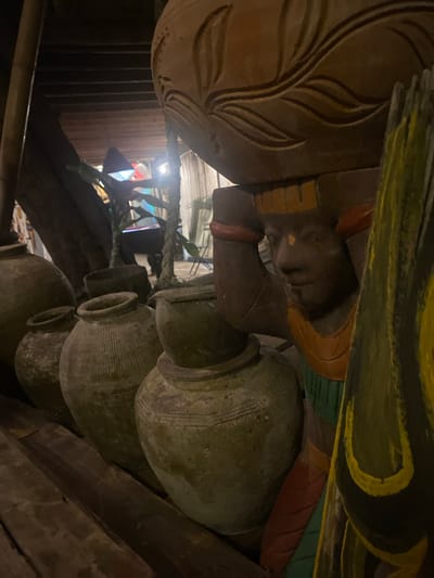 The image shows a close-up, upward-angle view of carved wooden figures and a stack of earthen pots in Baan Don Sai, Thailand. The figures appear to be part of a larger structure or sculpture, with one figure depicting a human face and body adorned with colored accents (yellow, green, red). The pots are stacked vertically, their surfaces textured and aged. A rope or vine is visible in the background, along with glimpses of an interior space, including wooden beams, corrugated metal, and colored lighting.