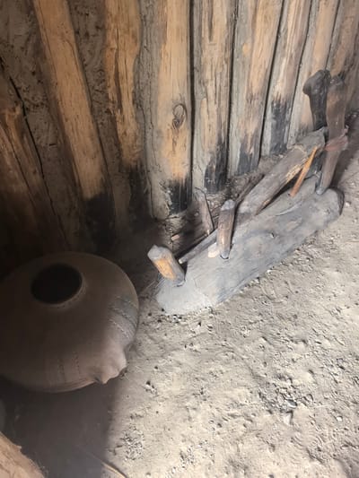 A wide, high-angle shot shows the interior of a rustic, wooden structure with a dirt floor. The walls are constructed from vertical, rough-hewn logs, some of which appear charred. On the left side of the frame sits a large, round, clay pot with a wide opening and decorative bands. To the right of the pot, a large, weathered wooden beam is positioned horizontally on the dirt floor, with several smaller wooden stakes or pieces of wood protruding from it and embedded in the ground. The ground is covered in sandy soil, scattered with small stones and debris, and appears to have several footprints. The lighting is dim and appears to be natural light filtering into the structure. This scene is located within the Камянюцкі сельскі Савет, Belarus.