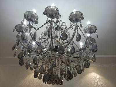 A luxurious crystal chandelier hangs from a white ceiling in a well-lit room. The chandelier has a metallic silver frame and is adorned with numerous faceted crystal teardrops and smaller beads. Eight light bulbs, emitting a warm, white light, are visible among the ornate branches. The crystals catch the light, creating a dazzling effect. The background shows a textured wall with a subtle pattern, and a decorative molding along the top of the wall. The scene suggests an interior setting, likely a home or a formal establishment, in Keles, Uzbekistan. The lighting indicates it could be daytime with artificial lights on, or evening. There is no text visible in the image.