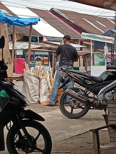 A man stands by a motorcycle at a roadside stall in Pulorayeuk, Indonesia. The man, wearing a black t-shirt and blue jeans, appears to be interacting with the stall owner. The stall sells sugar cane, ice, and what appears to be Indonesian street food. The scene is typical of a local marketplace, with vendors and motorcycles common.