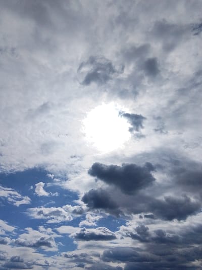 The image shows a cloudy sky in Treshchevka, Russia. The sun is obscured by a layer of clouds, appearing as a bright, diffuse circle in the upper center of the frame. Above the sun, the clouds are primarily gray and dense. In the lower portion of the image, the sky is a mix of blue patches and white, fluffy clouds. There is no evidence of people, subjects, objects, or actions.