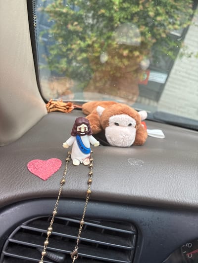 This image depicts items on the dashboard of a vehicle, likely in Balch Springs, United States of America. A small figurine of Jesus wearing a sash that reads "Jesus is Yo!" is positioned on the right side of the dashboard. To the left of the figurine is a small, pink, heart-shaped sticker. A beaded chain, possibly part of a rosary, drapes from the Jesus figurine. In the background, a plush toy monkey lies on its side. The dashboard is made of brown, textured material. A portion of the car's air vent is visible in the lower foreground. Through the windshield, green foliage and a building can be seen.