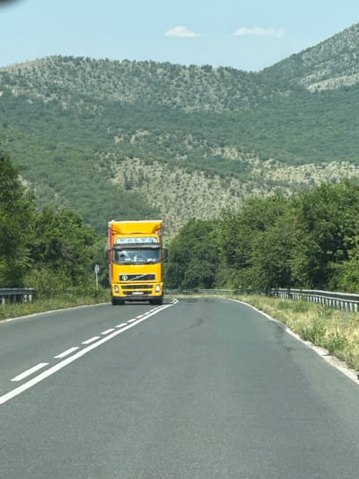This scene, located in the city of Binkos, Bulgaria, depicts a two-lane asphalt road traversing a mountainous landscape under a light blue sky. A bright yellow Volvo truck, displaying an 'L' sticker on its grille and a red side on its cargo unit, approaches the viewer. The road features white dashed double lines in its center and white solid lines on its shoulders, gently curving to the right. Metal guardrails are present on both sides of the road. Dense green deciduous trees line the roadside and cover the lower slopes of the background hills. The higher elevations of the hills exhibit sparser, lighter-colored vegetation and exposed rocky terrain. A few small white clouds are visible in the sky. No individuals are present in the image.