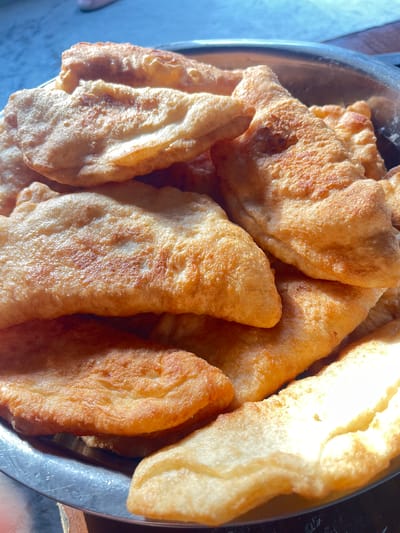 A close-up, overhead shot displays a silver bowl filled with freshly fried dough pieces, likely some form of pastries or fritters, commonly found in Central Asian cuisine. The dough is golden brown and appears slightly puffy, with some pieces showing a crispier texture. The lighting suggests it might be daytime, with a natural light source casting highlights on the food and the metallic sheen of the bowl.
The scene is likely set in a home or a casual eatery in Кызыл, Kyrgyzstan, given the provided location context. The background is slightly out of focus, but hints of a simple table setting or kitchen environment can be observed. There is no visible text in the image. The overall impression is one of simple, homemade food, ready to be served or enjoyed. The focus is entirely on the appetizing texture and color of the fried dough, evoking a sense of warmth and culinary tradition.