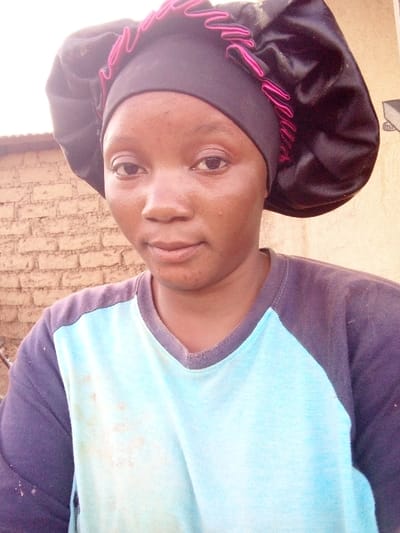 A medium close-up, eye-level portrait captures a person, likely a woman, looking directly at the camera. She has a dark complexion, dark eyes, and full lips, with a calm or slightly pensive expression. She appears to be in her late teens or twenties.
She is wearing a black sleep bonnet or hair cover with ruffled pink trim along the edge that frames her face. The main part of the bonnet, covering the back of her head, has a shiny, satin-like texture. Her outfit consists of a light blue short-sleeved or three-quarter-sleeved t-shirt with dark grey or black raglan sleeves and a V-neck collar. There are several faint brownish stains on the front of her light blue shirt.
The background behind her features a rustic, light brown brick or mud-brick wall on the left, suggesting an outdoor or semi-outdoor setting. On the right side, the background is lighter, possibly a different part of the wall or an area illuminated by sunlight. The lighting in the image suggests it was taken during the day, possibly outdoors due to the brick wall and bright illumination. No visible text is present in the image.