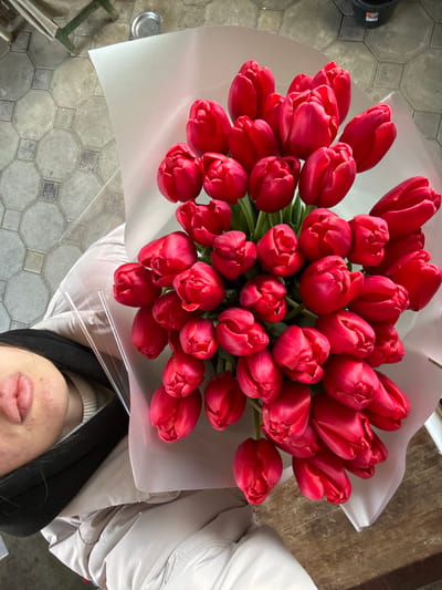 The scene is an interior view within Trudobelikovskiy, Russia, depicting a person holding a large bouquet of red tulips. The bouquet, comprising approximately 40-50 tightly arranged, vibrant red tulips, is wrapped in light-colored, semi-transparent paper and clear cellophane. The person's lower face, including the chin and full lips, is visible on the left side of the frame. The individual is dressed in a light-colored (beige or light grey) padded winter coat, with a dark inner garment and a light-colored high-neck shirt visible underneath. The background features a tiled floor with an octagonal pattern in shades of grey and beige on the left, and a wooden surface, possibly a table or counter, on the right. Distant background elements include a small metal container, a dark bucket, and a green-painted metal structure. The image is captured from an overhead perspective, with natural light illuminating the scene.
