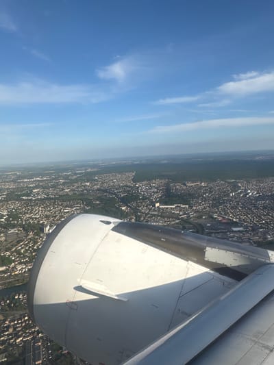 The image is taken from an airplane window, showing the wing of the plane and a view of the ground below. The sky is blue with some white clouds. Below, the ground is covered in dense urban development, punctuated by patches of green, possibly parks or forests. Given the prompt mentions Villeneuve-le-Roi, France, it is plausible this is the city from above. The wing of the plane is prominently featured in the right side of the frame. There are no people visible in the image.