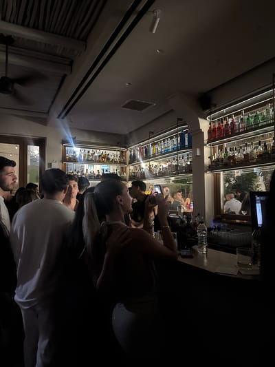 The image depicts an indoor scene at a bar in Naousa, Greece, characterized by dim lighting and a crowded atmosphere. Several individuals are present, with some gathered around a bar area. In the foreground, a woman with her back to the camera, wearing a light-colored top, is looking towards the bar and holding her phone up, possibly to take a photo or video. To her left, a man in a white t-shirt is also looking in the same direction. Behind them, a man with dark hair is visible. The bar itself is stocked with numerous bottles of alcohol on illuminated shelves, suggesting a well-appointed establishment. There are glasses and a bottle of water on the bar counter. In the background, the reflections in a mirror show other patrons and an outdoor scene with some greenery. The ceiling features a textured, possibly wooden, design and a ceiling fan. The overall impression is of a lively evening at a bar or nightclub.