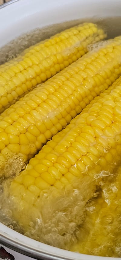In a white pot, three ears of corn are submerged in boiling water. The corn is bright yellow and appears to be fresh. Bubbles rise to the surface of the water, indicating that it is actively boiling. The image is a close-up shot, focusing on the corn and the water. The background is blurred, but it appears to be a kitchen or cooking environment. The scene is located in the city of Овощной, Russia.