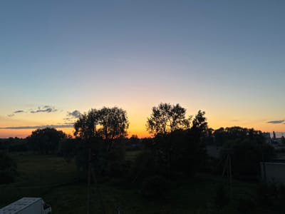 The image captures a twilight scene overlooking a landscape with a gradient sky transitioning from a clear, pale blue at the top to hues of orange and yellow near the horizon. Silhouetted trees and a line of buildings dominate the middle ground and background. In the foreground, a white van is partially visible on the left. The overall impression is one of a rural or semi-rural setting at dusk, possibly in the village of Maloye Verevo, Russia.