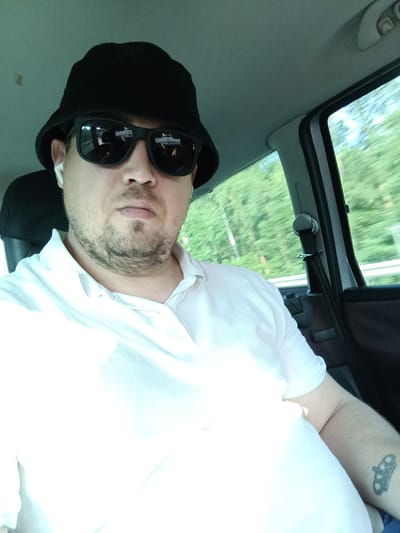A man is taking a selfie from inside a car. He is wearing a black bucket hat, black sunglasses, and a white polo shirt. He has short, light brown hair and stubble. He is also wearing white earbuds. The car appears to be in motion, as evidenced by the blurred green trees and foliage visible through the window. The sky is bright, suggesting it is daytime. A seatbelt is visible across the man's chest. On his left arm, a tattoo of a crown is visible. The location is Hvězdonice, Czechia.