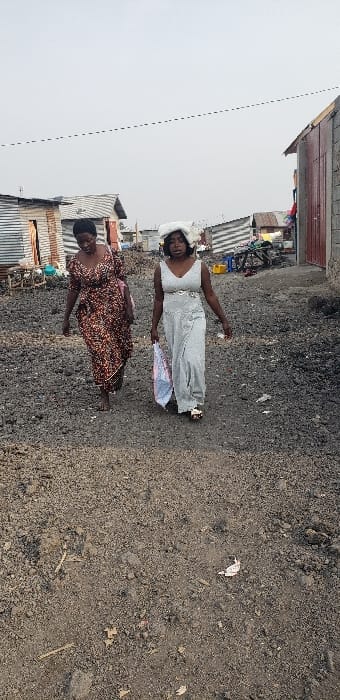 Two adult females are observed walking forward on an unpaved, dark earth path in Buzove, Democratic Republic of the Congo. The path is strewn with small debris and appears uneven. The woman on the left wears a long, patterned dress predominantly in shades of brown, red, and orange, and is barefoot. The woman on the right wears a long, light grey, possibly shimmering, sleeveless dress with a decorative belt; she wears light-colored sandals, carries a translucent plastic bag with pink and blue designs in her left hand, and balances a large, white, folded item on her head. In the background, multiple rudimentary structures, some constructed from corrugated metal sheets and others from concrete blocks, are visible under an overcast sky. A bicycle and a yellow jerrycan are identifiable among the background structures. A utility line extends horizontally across the upper portion of the frame.