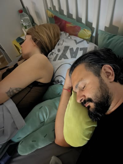 The image depicts a close-up view of a man and a woman sleeping in a bed. The man, on the right, has dark hair, a beard, and is resting his head on a light green pillow. His eyes are closed, and he appears to be asleep. He is wearing a black shirt. The woman, on the left, is lying on her side, facing away from the camera. Her hair is blonde, and she is wearing a black lace garment. She has a tattoo on her left arm. Various pillows are visible in the bed, including a white one with a gray pattern and a light green, cactus-shaped pillow. A bottle of water with a red cap is on a bedside table in the background, along with some colorful fabric. The scene is indoors, likely a bedroom, with a bed and bedding. The lighting suggests it is either nighttime or a dimly lit room. The location is identified as Mollet del Vallès, Spain.