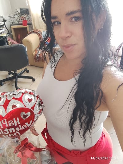 A woman with long, dark hair and blue eyes poses in a selfie. She wears a white tank top and red bottoms. A Valentine's Day balloon with red hearts and silver writing is partially visible. The date "14/02/2026" is printed at the bottom. A desk chair and other furniture are blurred in the background.
