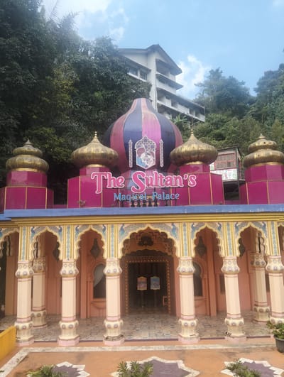 In the city of Gudangkahuripan, Indonesia, an outdoor shot captures the facade of "The Sultan's Magical Palace." The building features a vibrant, oriental-inspired design with prominent domes and ornate pillars. The main structure is painted in shades of pink and blue, with decorative gold accents on the domes and trim. Archways with intricate patterns, including peacock motifs, lead into the building. The entrance appears to be a dark opening, with signs displayed on stands in front of it. Lush green trees and a hillside form the background, with a multi-story building visible behind the palace's main structure. The sky above is partly cloudy. No people are visible in the image.