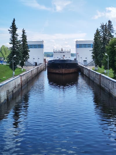 The image shows a view of the Uchinsky Water System's lock in Belousovo, Russia. A barge is positioned within the concrete lock canal, approaching a gate structure. On either side of the canal are concrete walls. In the background, two identical white buildings with blue accents flank the canal, and beyond them, trees and a body of water are visible under a partly cloudy sky. The overall scene depicts a functional industrial waterway infrastructure.