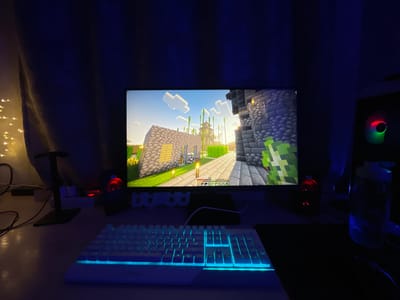 The image depicts a gaming setup in a dimly lit room. A computer monitor displays a scene from the video game Minecraft, featuring pixelated structures and characters. In front of the monitor is a white keyboard with blue backlighting. To the right of the keyboard is a black mousepad and a black computer mouse. Red LED lights from speakers are visible on either side of the monitor. A water bottle sits to the right of the setup. A white curtain with string lights is visible in the background to the left. The scene is set against a dark blue backdrop, suggesting ambient lighting from the computer. There is no discernible location in the city of Ардонь, Russia, or any people present.