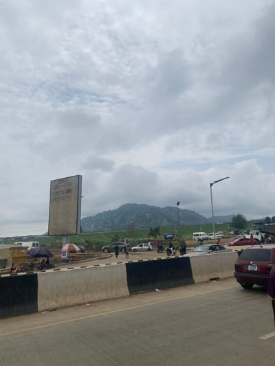 This is a full-shot, eye-level, outdoor photograph taken in Pasere, Nigeria, on an overcast day. The foreground features a highway with a concrete barrier separating it from the background. The barrier is painted black and white. Beyond the barrier, there is a busy scene with several vehicles, including cars, motorcycles, and a white van. People are seen walking and interacting.
To the left, a tall billboard with some writing on it is visible. The writing appears to be phone numbers: "CALL 08022671856" and "07035683958".
In the middle ground, there are some temporary stalls with umbrellas and goods being sold, suggesting a market or roadside vendors. A few people are gathered around these stalls.
The background features a lush green landscape with a prominent rocky hill or mountain. Several trees are scattered across the landscape. The sky is filled with dense, grey clouds, indicating a possibility of rain. The overall impression is a bustling roadside scene in a semi-urban or rural area with a distinct geographical feature in the background.
