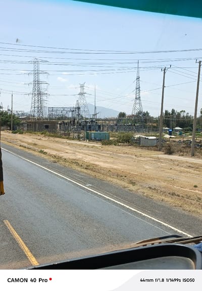 The image displays an outdoor daytime scene in Hells Gate ward, Kenya, captured from within a vehicle. In the foreground, a dark dashboard and a brown windshield wiper blade are visible at the bottom edge. An asphalt road with faded white and yellow lane markings extends from the lower left towards the center. Adjacent to the road, a wide expanse of dry, light brown soil with sparse, dry vegetation occupies the midground.
The midground and background are dominated by extensive electrical infrastructure. Multiple lattice-steel transmission towers of various heights and designs, supporting numerous overhead power lines, are prominent. A large power substation is visible, containing several steel frameworks, insulators, transformers, and switchgear. Adjacent to the substation, a rectangular, single-story building with an unfinished, rough exterior and an empty window opening is present. Several container-like structures, including a green one and a smaller corrugated metal shed, are also within the substation area. On the right, wooden utility poles with cross-arms and lines are visible, along with some taller trees and distant, indistinct structures.
The background features a clear, light blue sky with a few wispy white clouds. Distant, hazy blue mountains or hills are visible along the horizon behind the electrical installations. No individuals, specific actions, or interactions are discernible within the frame.