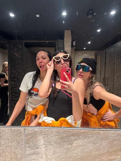 Three women pose in a mirror selfie. One wears a white t-shirt, and playfully sticks her tongue out while pointing up. Another holds a pink phone with beige sunglasses and wears a black shirt. The third, situated at the top, sports a black beanie and colorful sunglasses. An orange towel wraps around them. The dark room has shiny black tile with visible light reflections. A fourth person is vaguely seen at the bottom of the mirror.