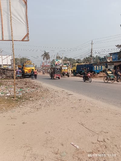 The image depicts a roadside scene in Kaliganj, Bangladesh, recorded on March 12, 2025, at 11:01. A two-lane paved road extends into the distance under a clear, bright sky. The road is flanked by unpaved shoulders.
On the left foreground, a bamboo pole structure supports a large, empty white rectangular banner frame. Below this, the ground is covered with scattered litter and debris. Further down the left side of the road, multiple yellow dump trucks are visible, one prominently in the mid-ground. Several individuals, including two people in dark clothing, are walking near the front of the closest yellow truck. An auto-rickshaw with a dark canopy is moving away from the viewer on the left lane.
On the right side of the road, an individual on a motorcycle is driving away from the viewer, while another person on a bicycle rickshaw is also moving in the same direction further along the road. A blue dump truck is parked on the right shoulder. Utility poles with numerous power lines crisscross overhead. Buildings and various billboards, including one with a portrait, line both sides of the road in the mid-ground and background. Palm trees and other foliage are visible against the skyline. The unpaved right shoulder shows tire tracks and minimal debris.